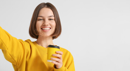 Beautiful woman with cup of coffee taking selfie on light background