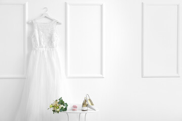 Stylish shoes, rings, bouquet of flowers on table and wedding dress hanging on white wall