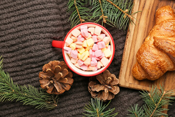 Cup of cocoa with marshmallows, fir cones, branches and croissant on warm scarf