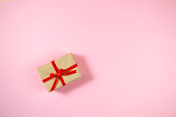 Holiday gift box wrapped in craft paper and tied with red ribbon on light pink background. Copy space in center. Flat lay. Copy space. Concept of holidays and gifts preparation.