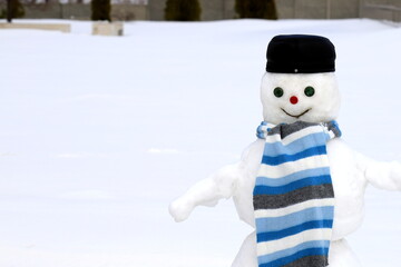 A funny snowman stands on snow covered playground in winter among white snow. A snowman in black cap and blue scarf, Christmas and New Year holidays, children entertainment and sports,