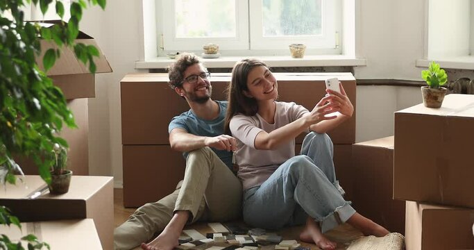Overjoyed young family couple first time apartment buyers making selfie with keys in hands on mobile phone among boxes with stuff. Happy millennial husband wife streaming live from new home using cell