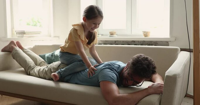 Laughing young daddy lying on stomach on sofa shaking moving forward. Little daughter sit on dad back imagine riding on fantasy dragon. Father and child girl play funny game