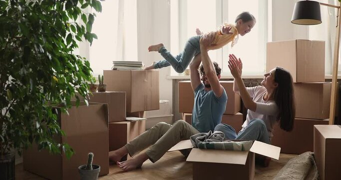 Playful little girl run to happy parents unpacking boxes at living room at relocation day. Excited father lift daughter on arms up to air while mother is laughing. Family celebrate moving in new flat