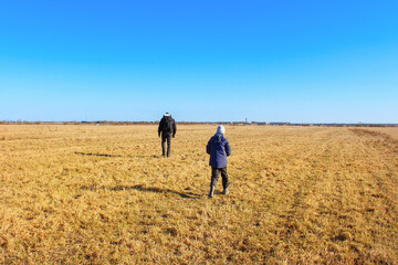Boy and man walking in field of dry grass under blue sky, view from the back, sunny day