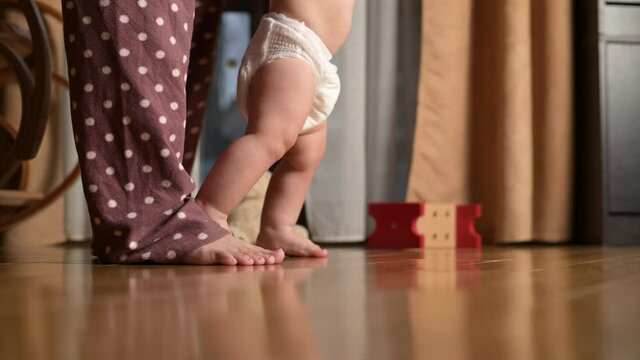 The first steps of a little boy around the house. The baby in the diaper learns to walk with the help of the mother. Close-up of legs