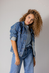 young blue-eyed blonde woman with curly hair in a denim jacket and jeans stands on a gray background. concept of fashion and beauty in the studio. beautiful clean healthy skin. space for text