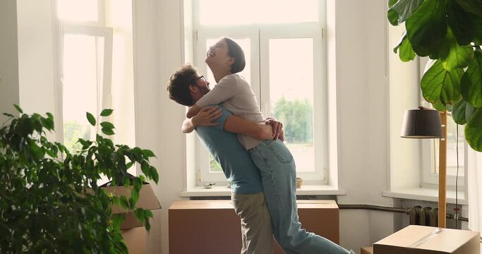 Laughing young husband cuddle raise up wife dangling feet in air near boxes with stuff overjoyed with moving to new apartment. Excited married couple feel happy of achieving goal getting mortgage loan