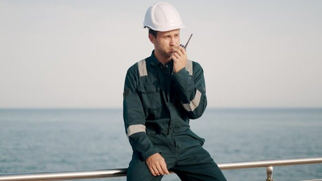 Young port worker in helmet and coverall using vhf radio, controlling work process in shipping port