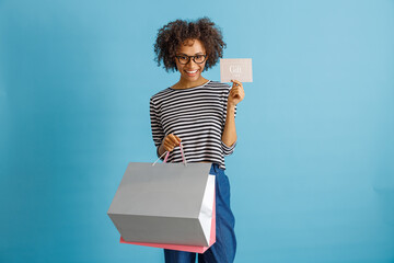 Cheerful Afro American lady looking at camera and smiling while holding shopping bags and gift certificate. Isolated on blue background