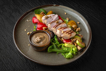 Grilled chicken steak with vegetables and sauce in a ceramic plate on a dark textured background. Restaurant menu Isolated on black