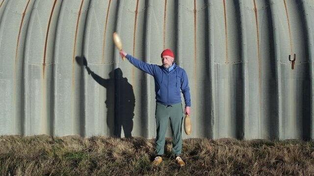 senior athletic male is swinging wooden Indian clubs with a company of a shadow on a grunge metal hangar, functional fitness workout in urban or industrial environment
