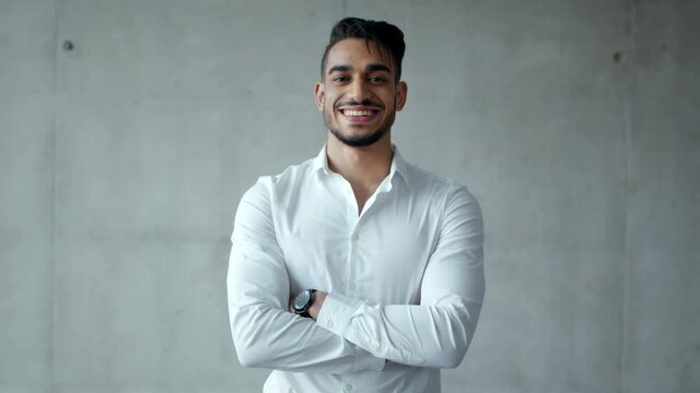 Young confident arabic businessman in formal shirt smiling to camera, posing with crossed hands, grey wall, slow motion