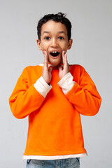 Funny mixed race kid boy in orange sweater posing emotionally on gray background