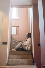 Pensive woman sitting on stairs at home cottage with hot beverage dreaming looking into window