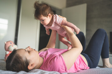 Young Adult caucasian mother playing with her four months old baby daughter while lying on the bed in bedroom at home parenting and motherhood family and happiness love concept