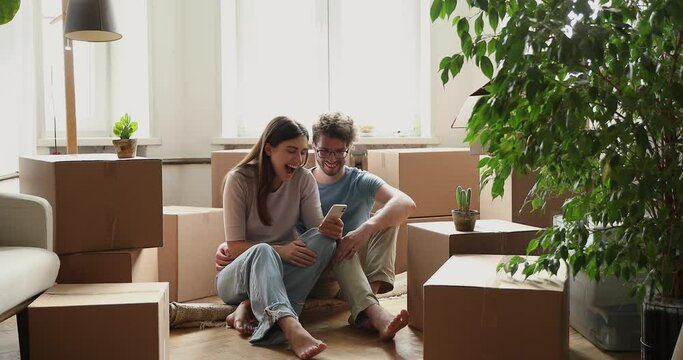 Enthusiastic spouses relax on floor near boxes at relocation day look on cell plan home improvement. Young couple flat buyers take break in unpacking belongings use phone laugh on funny design ideas