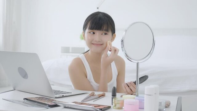 Beauty of young asian woman with learning makeup with powder puff on cheek watching laptop computer with tutorial course online, female teaching and explain make up with cosmetic on streaming.