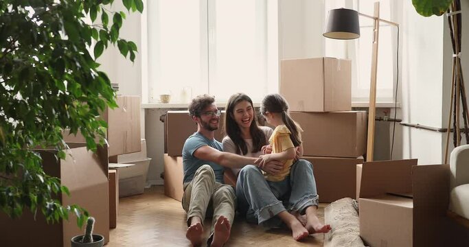 Joyful young foster parents little adopted daughter tickle laugh at empty living room at relocation day. Excited family new home owners have fun play on warm floor take break in unpacking belongings