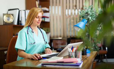 Professional smiling female doctor sitting in office at workplace and working at laptop