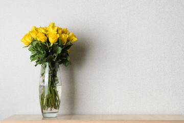 A large beech of yellow roses in a glass vase on a wooden shelf on a gray background, with a copy...