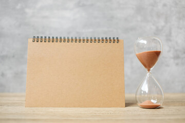 Hourglass with Blank notebook on table, Sand flowing through the bulb of Sandglass measuring the...