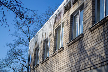 Fototapeta na wymiar Corner of brick house with icicles on the roof. Long, shiny icicles on bright sunny day. Frosty morning. Dangerous place.