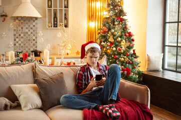 Teenage boy sits on the couch wearing a santa hat and with a phone in his hands. Online communication at Christmas. Holiday online shopping. Christmas atmosphere and relaxation.