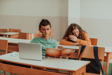 A boy sitting in a school classroom holding a book in his hand and uses a laptop. Selective focus
