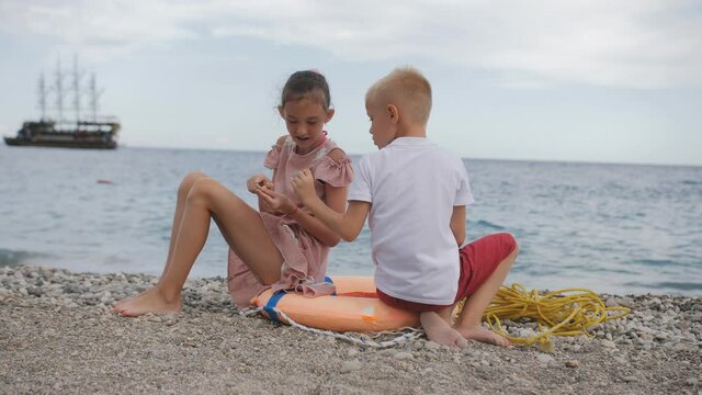 A girl and a boy of 8 years old sit together on a lifebuoy on the seashore and look at sea pebbles