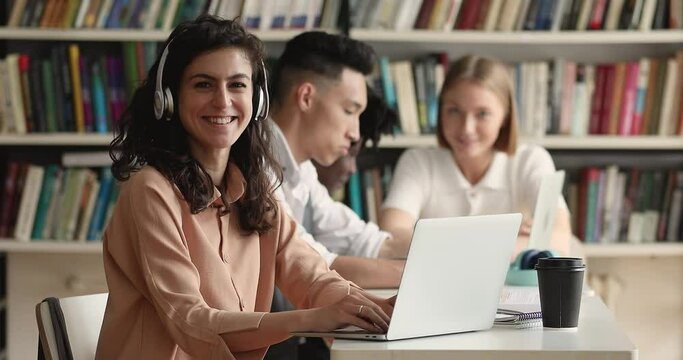 Portrait of Hispanic attractive student girl wear headphones studying sit at table in library with laptop, smile look at camera. Education, gain new knowledge use modern wireless tech, e-learn concept