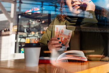 Clsoe up portrait of woman is talking on the phone and holding cash in her hands. View through the cafe window. The concept of business and freelancing