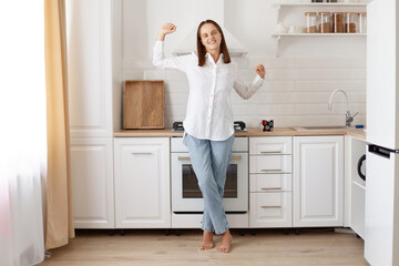 Indoor shot of young adult attractive woman wearing white shirt and jeans, standing at home against light kitchen set and stretching hands, yawning, looks sleepy.