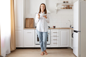 Positive optimistic female with dark hair and pleasant appearance wearing white shirt and jeans standing with plate in hands and eating soup, posing against kitchen set at home.