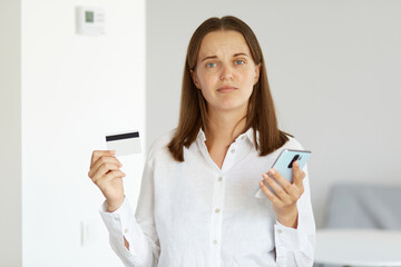 Fototapeta na wymiar Portrait of dissatisfied woman wearing white shirt standing indoor in light room, holding mobile phone and credit card in hands, looking at camera, online shopping, cashback.