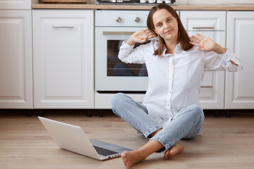 Indoor shot of tired woman sitting on floor in front of laptop computer and working, stretching her arms, looks sleepy, freelancer posing against kitchen set.