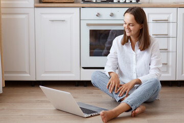 Portrait of beautiful woman sitting on floor with crossed legs in kitchen, looking at laptop display, wearing white shirt and jeans, having pleasant online conversation.