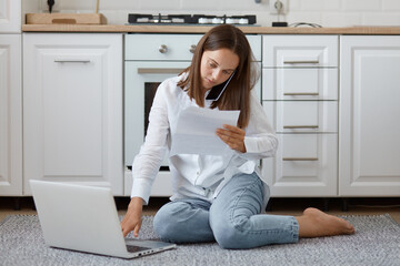 Portrait of serious concentrated Caucasian dark haired woman wearing jeans and shirt sitting against white kitchen set on floor, talking with utility company, holding bills in hands.