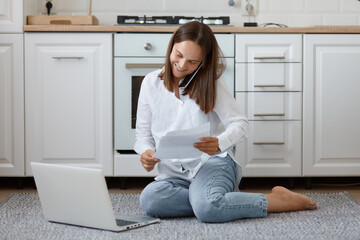 Smiling beautiful woman of young age sitting on floor in kitchen and talking phone, using laptop...