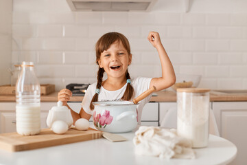 Fototapeta na wymiar Excited amazed little girl with pigtails posing in kitchen with products for baking cake or pie, having great mood, clenching fist and yelling happily with excitement.