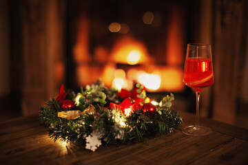 Christmas wreath, glass of champagne, blurred background burning fireplace with fire. Warm cozy Xmas or new year holiday evening home