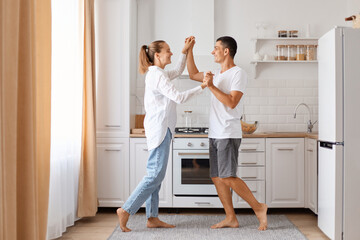 Obraz premium Indoor shot of happy beautiful couple smiling while dancing in kitchen at home, holding each other hands, enjoying spending time together, family wearing white shirts and jeans.
