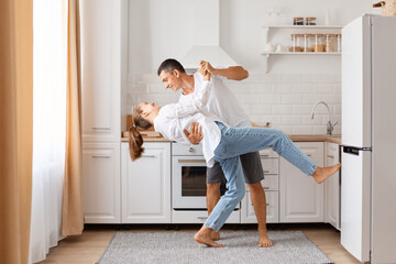 Side view full length portrait of happy excited couple dancing in kitchen in the morning, having fun together, enjoying weekend, posing at home, expressing romantic feelings.