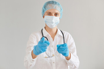 Indoor shot of serious confident doctor woman wearing medical cap, gloves, surgical mask and gown, standing looking and pointing to camera, choosing you for medical examination.