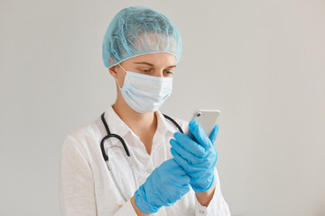 Indoor shot of concentrated female doctor wearing medical cap, gloves, surgical mask and gown, standing and using mobile phone, checking e mail or calling to someone.