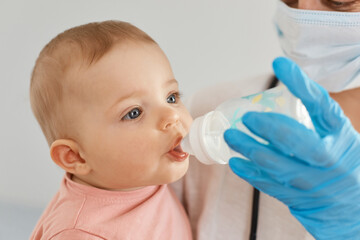 Indoor shot of unknown female doctor or nurse feeding baby girl from bottle, giving water to toddler child, woman wearing blue medical rubber gloves.