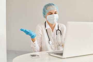 Confused female doctor wearing medical cap, surgical mask, rubber gloves and gown, using notebook...