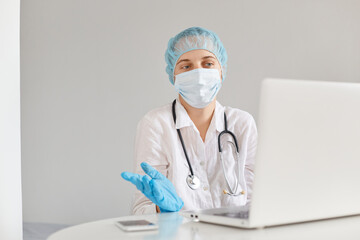 Confused puzzled woman doctor sitting at table and working on laptop computer, wearing medical cap, surgical mask, rubber gloves and gown, having confused expression.