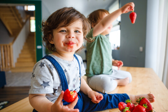 Little brothers eat strawberries are sitting on a wooden table in the kitchen.