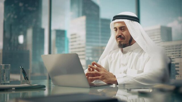 Successful Arab Businessman in White Traditional Outfit Sitting in Office and Working on Laptop Computer. Business Manager Planning Corporate Strategy. Saudi, Emirati, Arab Businessman Concept.